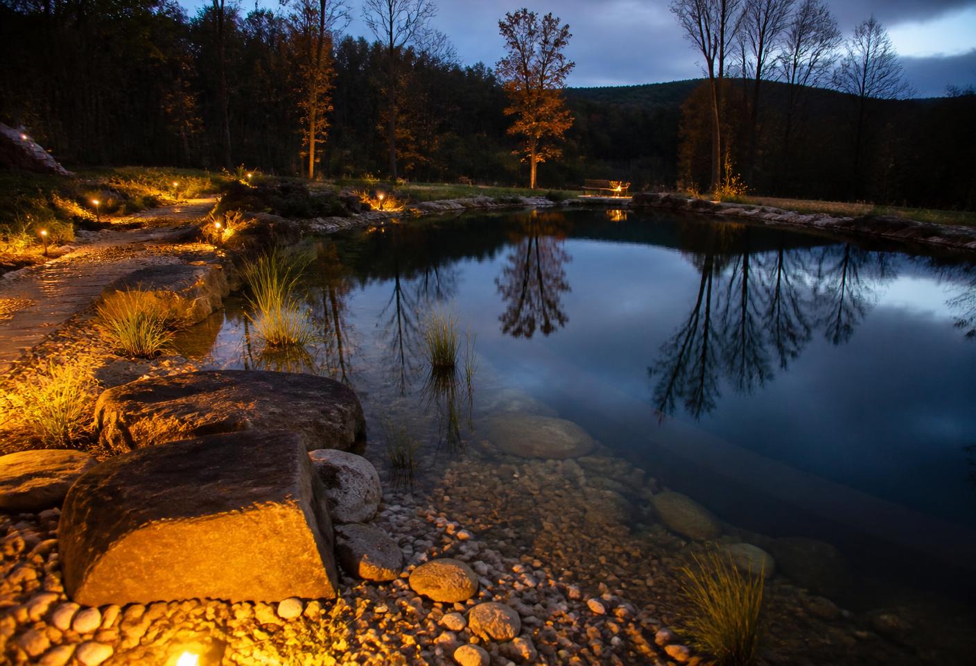 Natural swimming pond at dusk with warm path lighting and boulder edging